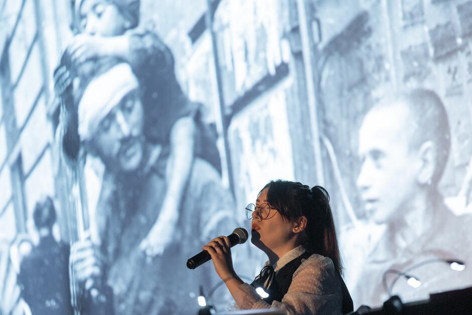 Kobieta śpiewająca do mikrofonu na tle biało-czarnego zdjęcia z czasów wojny
