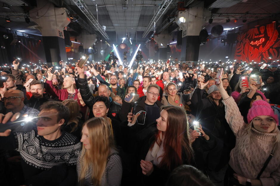 Tłum ludzi w sali koncertowej, w dłonaich niektórzy trzymają świecące telefony komórkowe