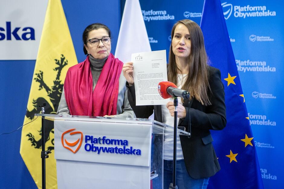 dwie kobiety na tle logo partii stają za mównicą, młodsza, długowłosa pokazuje dokument