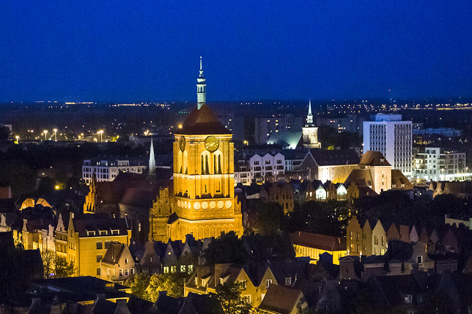 nocne zdjęcie miasta z lotu ptaka. Na pierwszym planie oświetlona wieża kościoła otoczona licznymi niższymi budynkami.