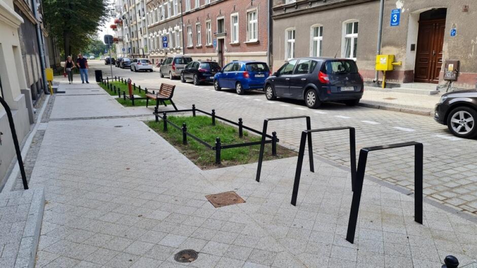 na zdjęciu wyremontowany chodnik, po prawej zamontowane trzy stojaki rowerowe, w tle widać trawnik, a także fragment ulicy i zaparkowane na niej samochody