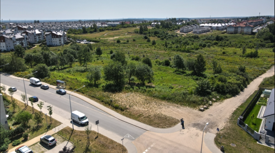 na zdjęciu z drona widać głównie zielony teren, porośnięty trawą i kilkoma drzewami, przed nim widać fragment jezdni, a w tle zabudowę mieszkaniową, niewysoką