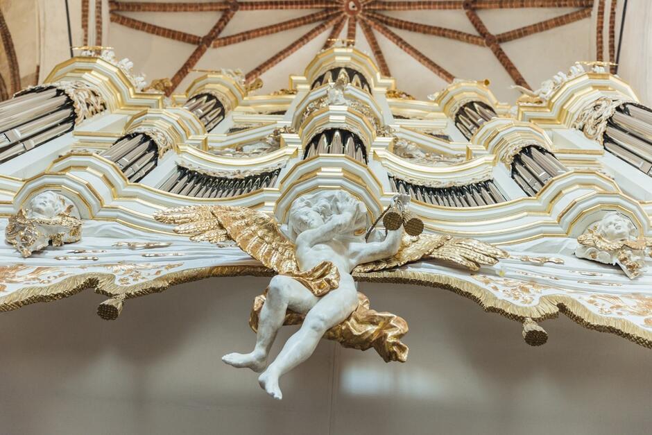 ogromna, biała konstrukcji szafy organowej, na pierwszym planie złota rzeźba anioła, nad nim piszczałki organów