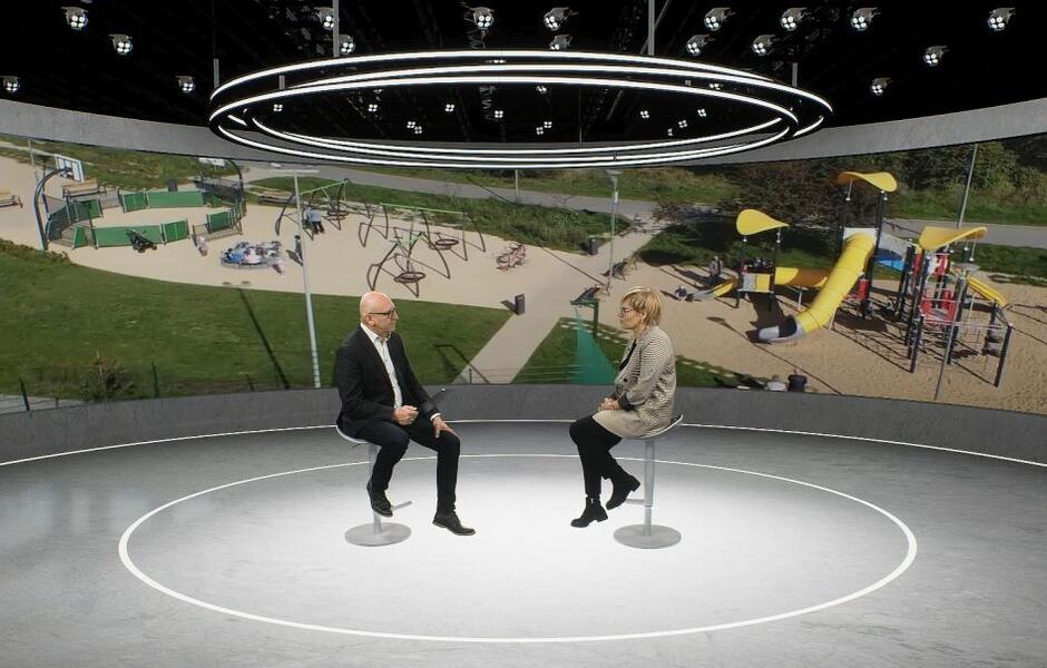 dwie osoby kobieta i mężczyzna siedzą i rozmawiają w wirtualnym studiu, w tle widać reprojekcję placu zabaw