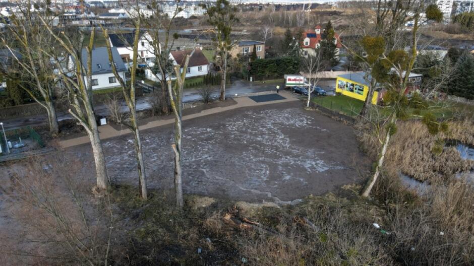 na zdjęciu z drona widać gruntowy teren z kilkoma drzewami, teren jest wyrównany, w tle po lewej widać zabudowę jednorodzinną