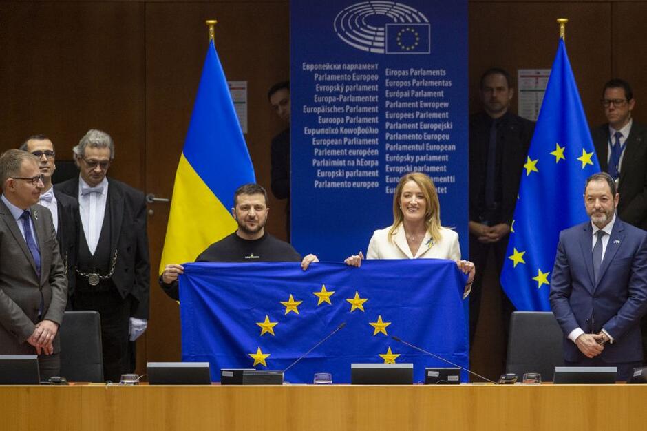 Pośrodku zdjęcia stoją mężczyzna i kobieta (z prawej) za stołem prezydialnym. Wspólnie pozują do zdjęcia z flagą Unii Europejskiej, którą razem trzymają w rękach. Po prawej i lewej, na marginesach zdjęcia widać kilka innych osób