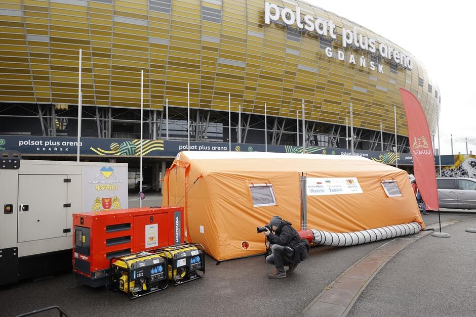 Na placu stoi duży podłużny pomarańczowy namiot, obok niego jeden większy generator prądu i dwa mniejsze. Widać fotoreportera, który robi zdjęcia. W tle znajduje się bryła stadionu Polsat Plus Arena Gdańsk