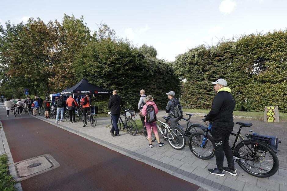 na zdjęciu kilkunastu rowerzystów, stoją w kolejce, jeden za drugim, z rowerami, w tle widać niewielki czarny namiot, jest też fragment chodnika i drogi rowerowej