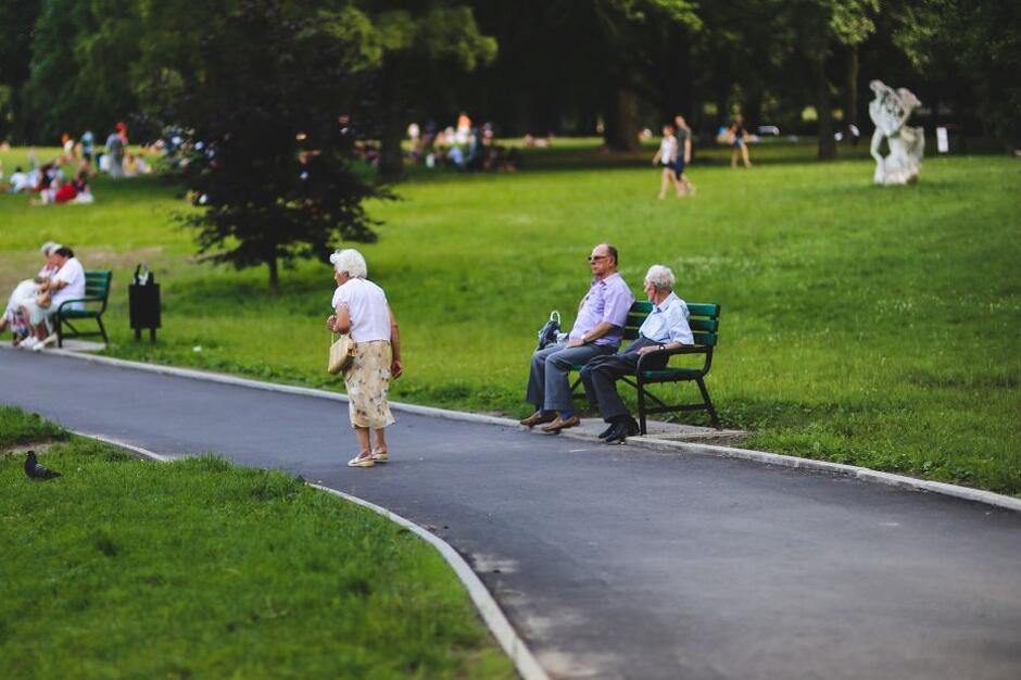 seniorzy w parku, alejką spacerują staruszka, a starsi panowie dwaj siedzą na ławeczce