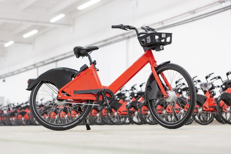 Na pierwszym planie czerwony rower miejski, za nim widoczne rzędy takich samych rowerów ustawione w hali