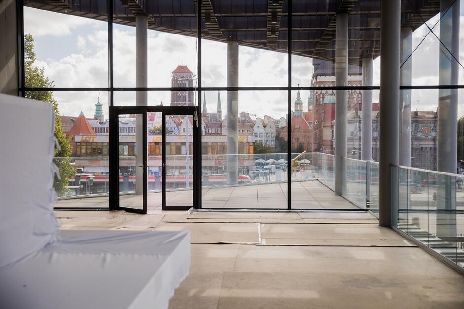 zdjęcie wykonane w budowanym pomieszczeniu, widać ogromne okno, a za nim zabytkowego budynki głównego miasta Gdańska 
