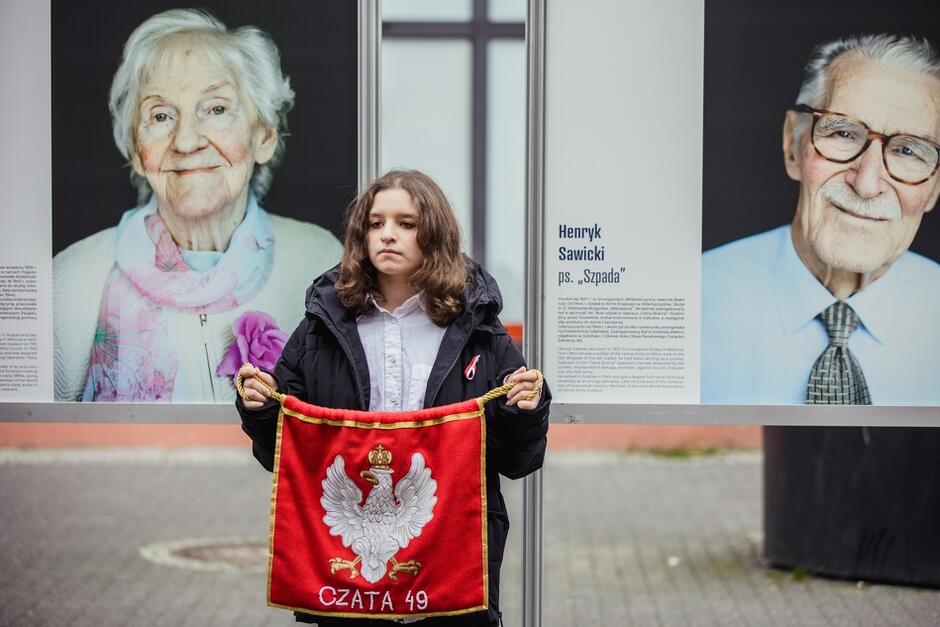 Zdjęcie przedstawia dwa uliczne fotosy z portretami weteranów AK. Zofia Zygmuntowicz widoczna jest po lewej, po prawej widzimy portret mężczyzny. Przed fotosami stoi młoda dziewczyna, która trzyma czerwoną chorągiewkę z białym orzełkiem w koronie, pod którym jest napis CZATA 49 