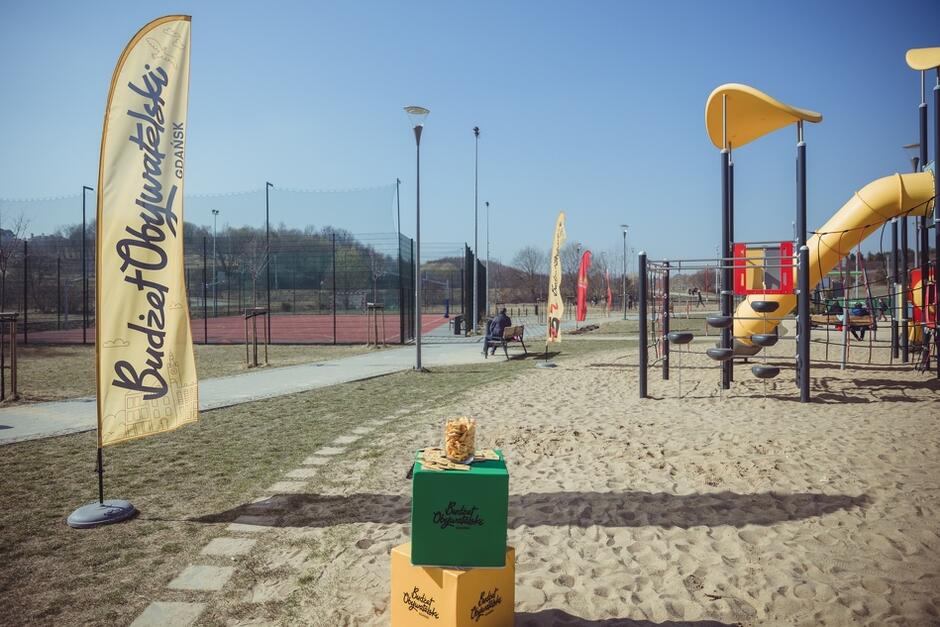 na zdjęciu fragment nowego placu zabaw, widać kolorowe urządzenia, po lewej wysoka i wąska żółta flaga z napisem budżet obywatelski