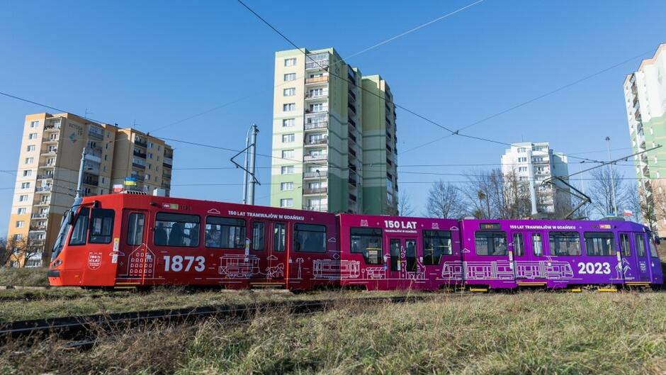 Kolorowy tramwaj stoi na torach, za nim długi blok mieszkalny 