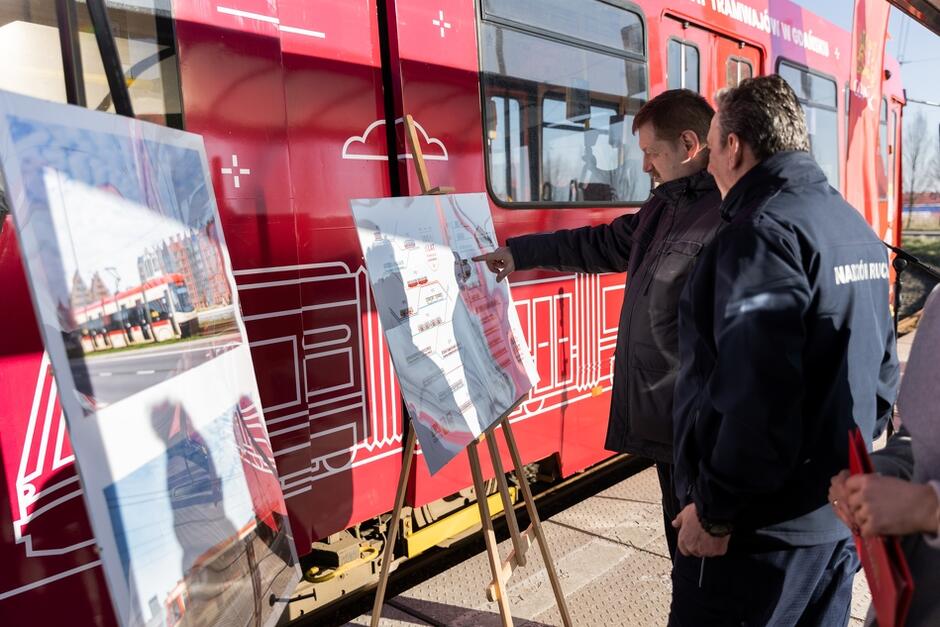 Dwaj mężczyźni oglądają wystawę zdjęć z historycznymi tramwajami. Za stojakami ze zdjęciami stoi tramwaj 