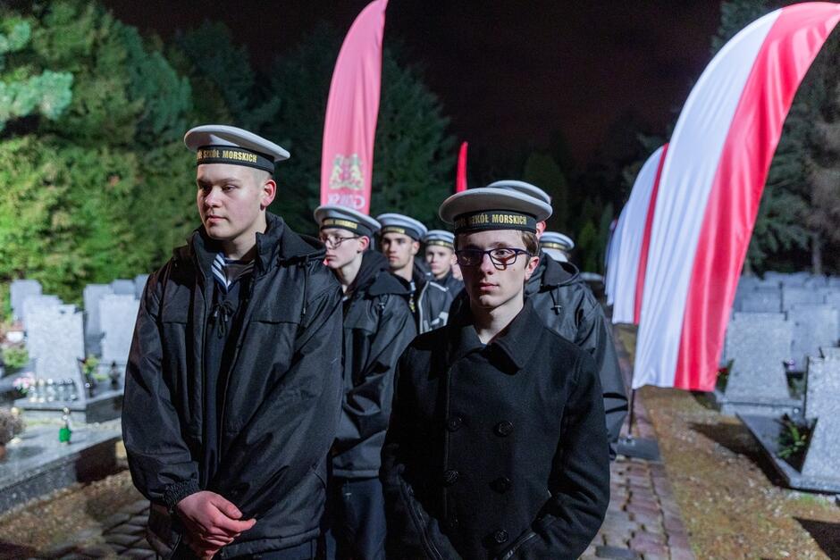 uczniowie w strojach szkolnych - mundurki i czapki stylizowane na marynarskie stoją nocą w cmentarnej scenerii