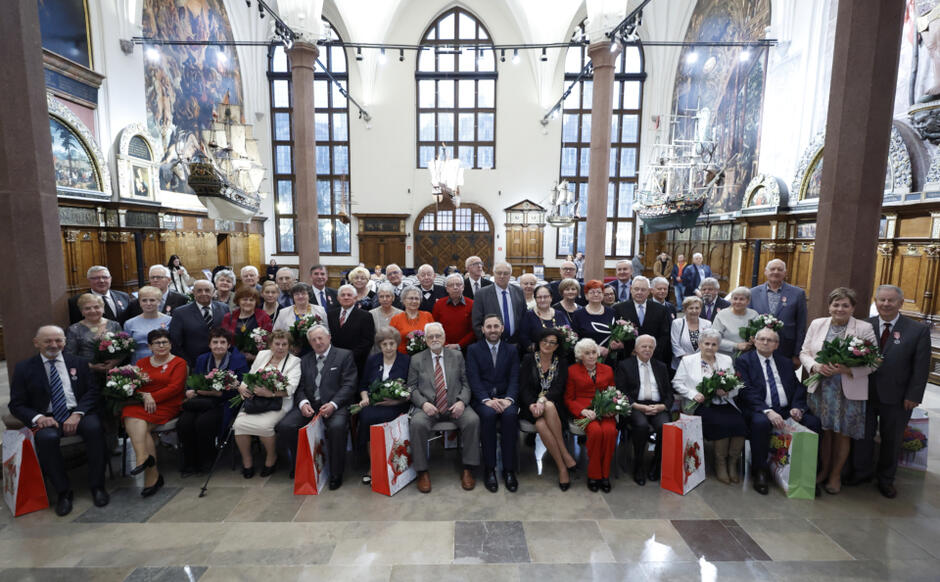 25 par odświętnie ubranych jubilatów pozuje do zdjęcia pamiątkowego w zabytkowych wnętrzach