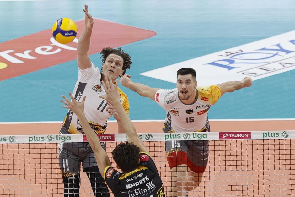 Mecz siatkówki: za siatką dwóch zawodników w białych koszulkach, przed siatką w czarnej koszulce zawodnik z wyciągniętymi do góry rękoma 