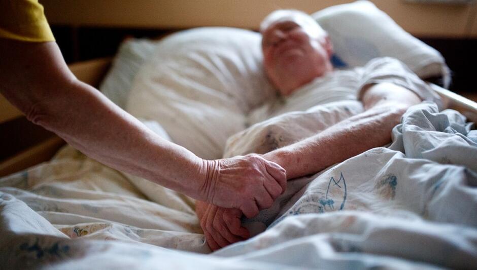 Osoba starsza leży w łóżku, obok niej z lewej strony widać rękę trzymają za rękę osobę leżącą w łóżku