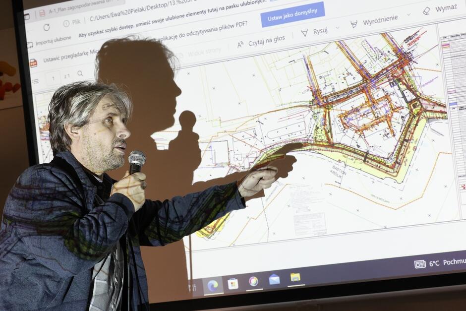 na zdjęciu mężczyzna, trzyma mikrofon w jednej ręce, w drugą pokazuje na wyświetloną mapkę