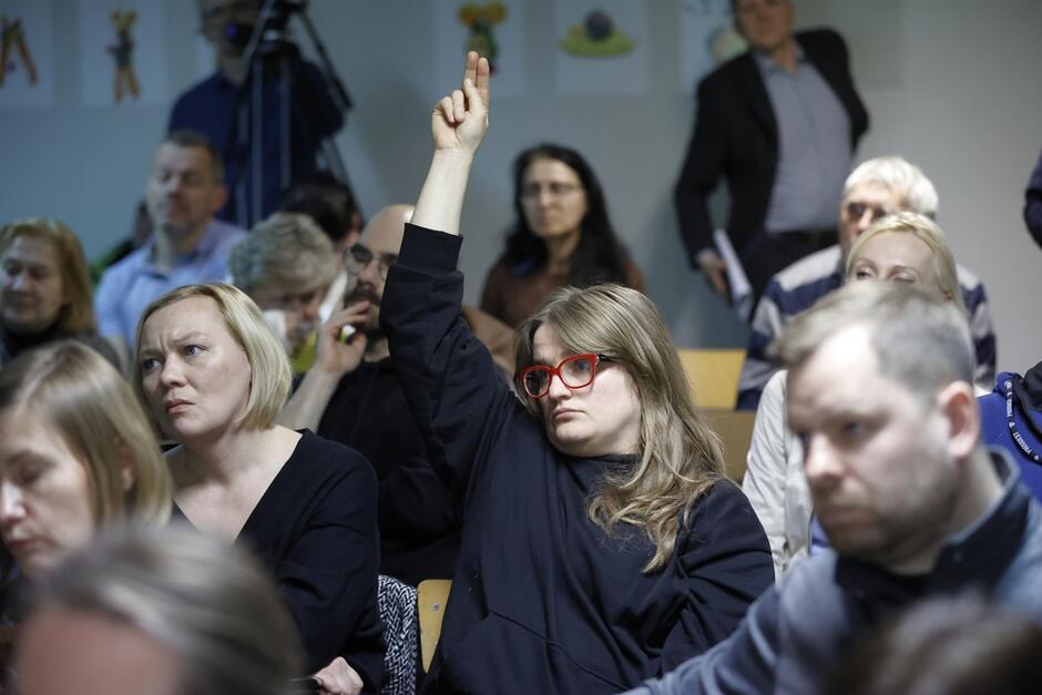 na zdjęciu młoda kobieta w czerwonych okularach, siedzi, podnosi rękę zgłaszając chęć zabrania głosu, wokół niej siedzi kilka innych osób