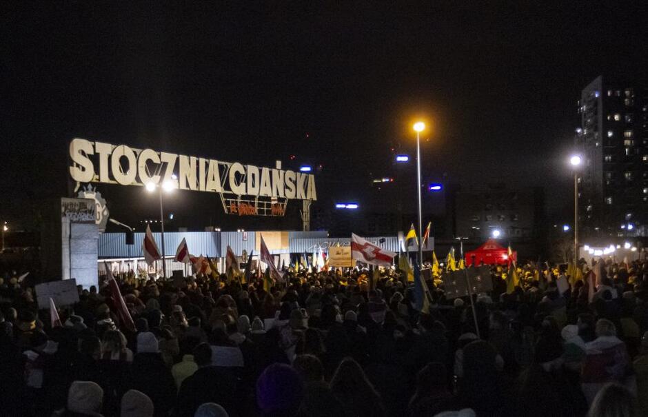 tłum ludzi pod nocą, stoją pod bramą z napisem Stocznia Gdańska