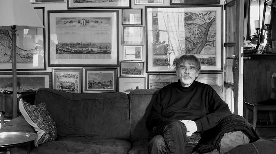 Gunnar Heinsohn siedzi na kanapie w dolnej części zdjęcia. Powyżej jest duża ściana gęsto zawieszona mapami i rycinami w ramach 