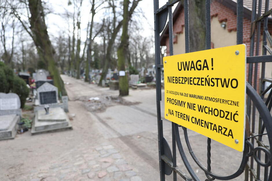 Brama cmentarza uchylona, na niej żółta tablica z napisem: UWAGA NIEBEZPIECZEŃSTWO. ZE WZGLĘDU NA ZŁE WARUNKI ATMOSFERYCZNE PROSIMY NIE WCHODZIĆ NA TEREN CMENTARZA