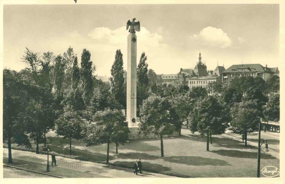 Wysoki, pionowy pomnik (obelisk) wykonany z białego kamienia. Na górze czarny niemiecki orzeł, tzw. gapa. Pomnik stoi w otoczeniu zieleni. W oddali zabudowa dalszych ulic.