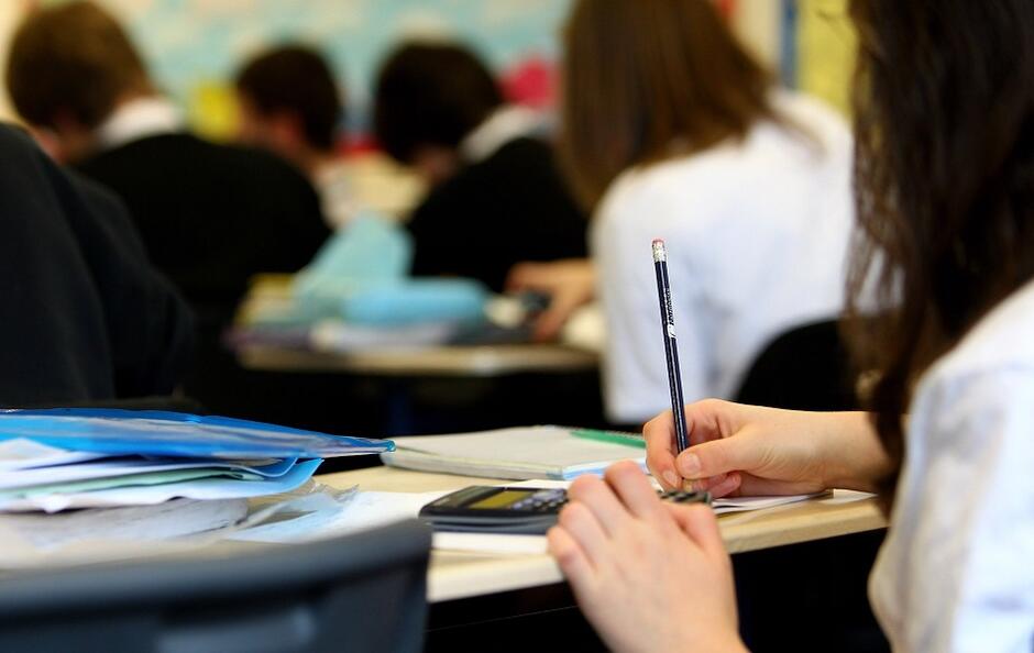 sala szkolna, plecy dziewczyny siedzącej przy ławce, na stole kalkulator, w ręku ołówek