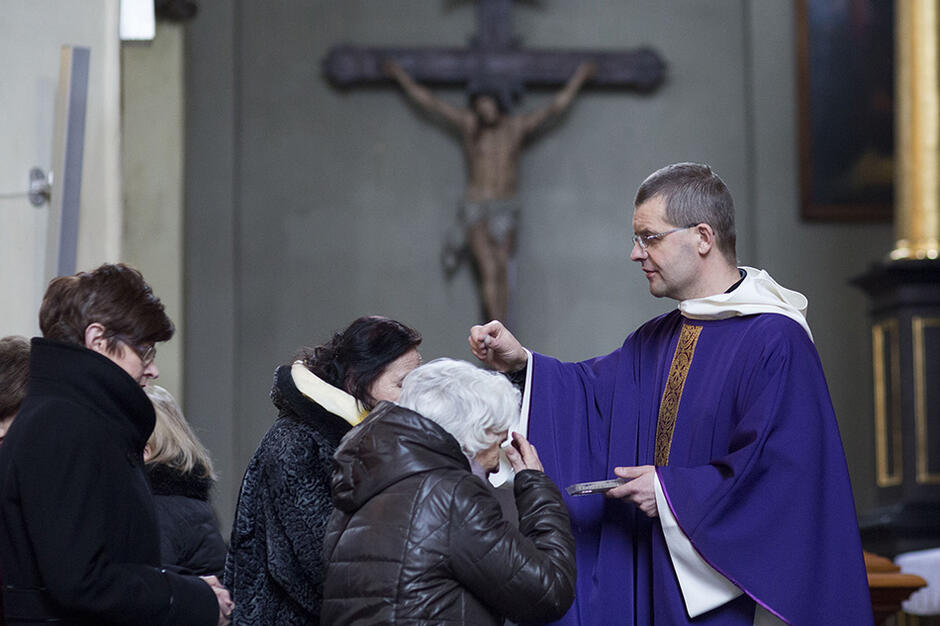 Zdjęcie przedstawia księdza, który soi po lewej, posypując kolejno głowy osób popiołem - osoby te sa ustawione gęsiego po lewej i po kolei podchodzą do duchownego