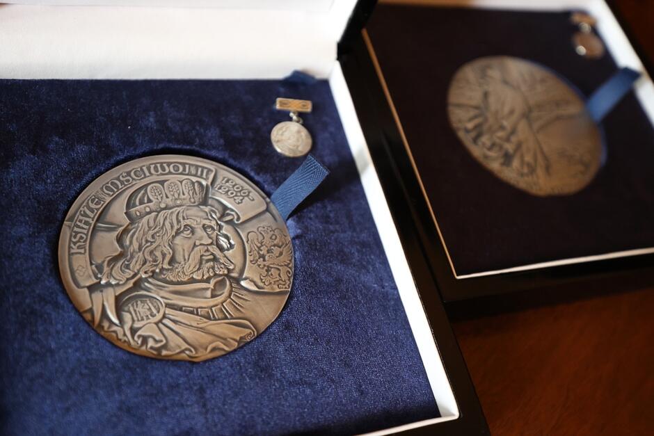 Awersy medali Księcia Mściwoja II (na pierwszym planie) oraz św. Wojciecha (mniej wyraźny, na drugim planie), ułożonych w etui.