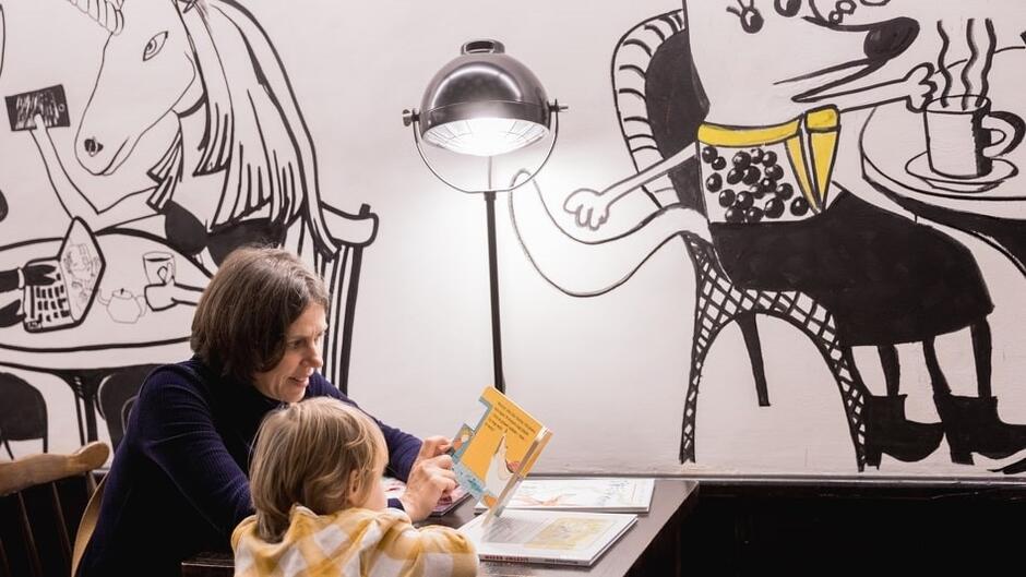 kobieta z dziewczynką siedzą przy stole przystawionym do ściany. Na ścianie kolorowy mural
