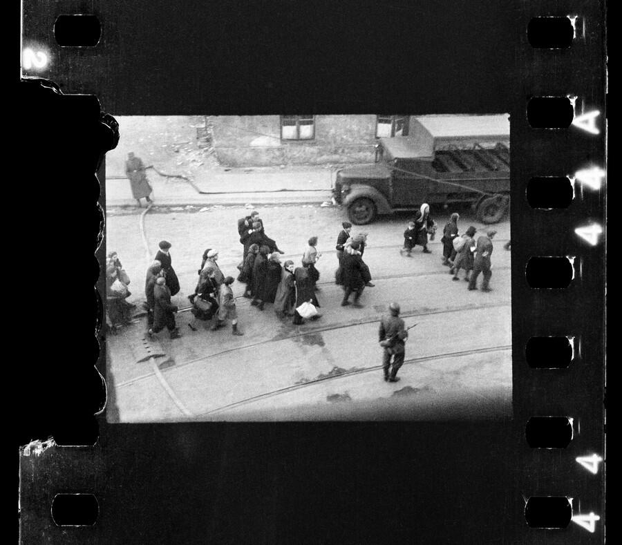 Czarno-białe zdjęcie, widok z góry: ulica, tory tramwajowe, idący środkiem ludzie z tobołkami, po bokach niemieccy żołnierze z karabinami. Powyżej samochód ciężarowy 