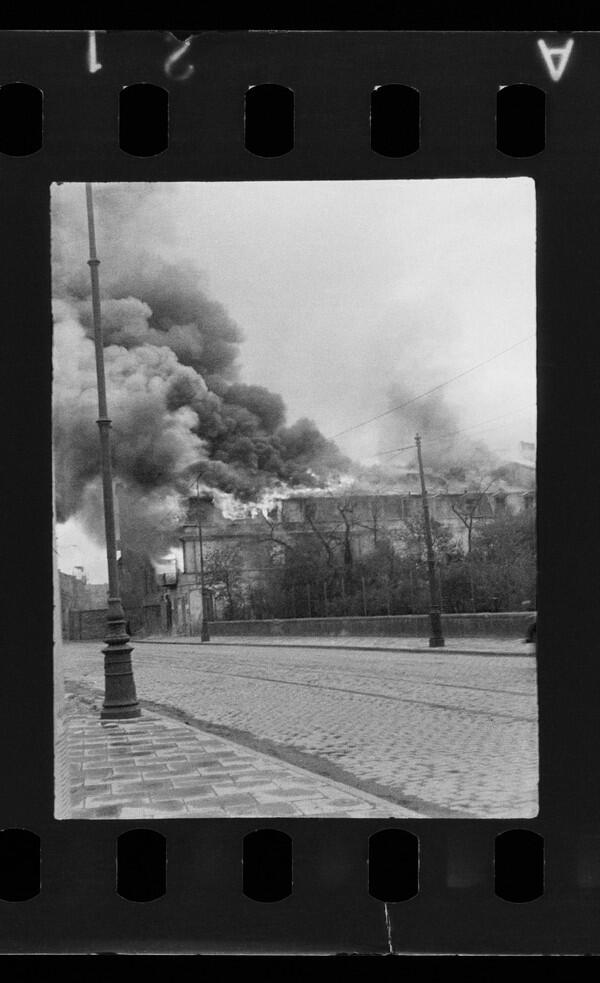 Czarno białe zdjęcie: ulica, na chodnikach latarnie, w tle piętrowy budynek, pali się dach, kłęby dymu