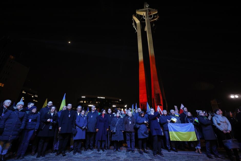 na zdjęciu widać tłum stojących osób, po prawej ktoś trzyma ukraińską flagę, w tle pomnik poległych stoczniowców.