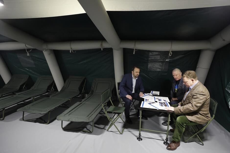 trzech mężczyzna siedzi przy stole zasłanym ulotkami w kolorach Ukrainy, są w namiocie, wokół stoją łóżka polowe