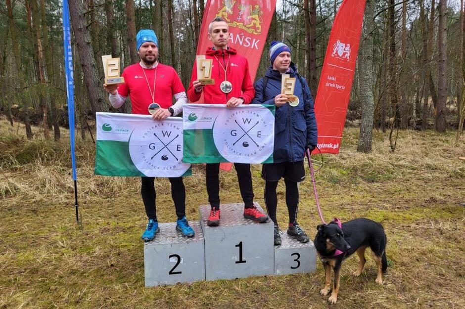 trzech mężczyzn z medalami stoi na podium ustawionym w lesie, obok pies