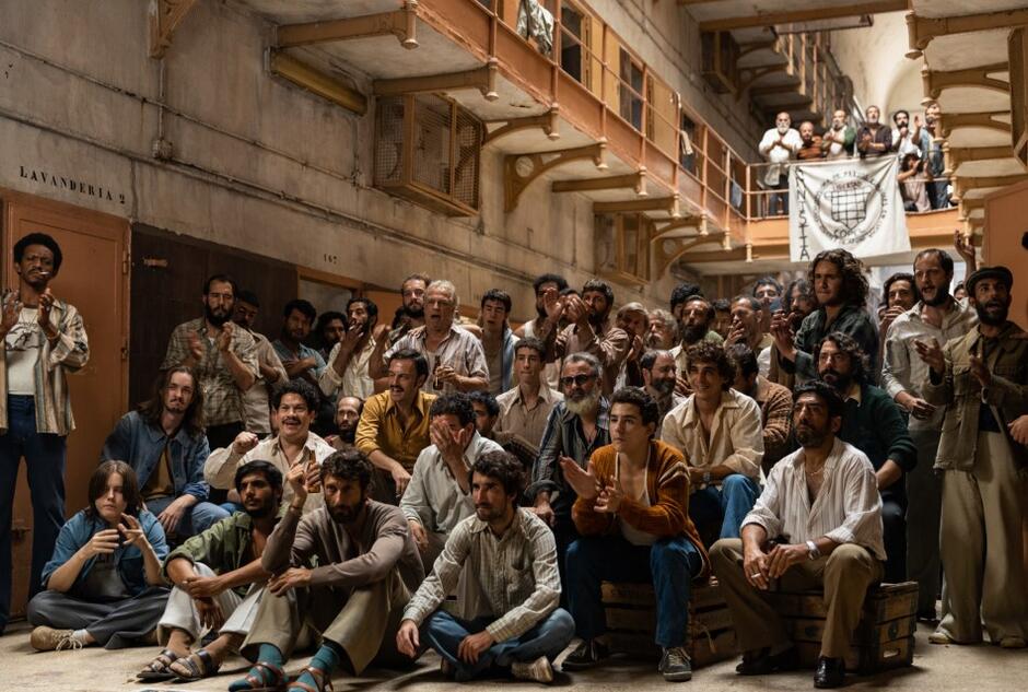 Un grupo muy grande de hombres con ropa de prisión sentados en las escaleras, en el espacio industrial circundante. 