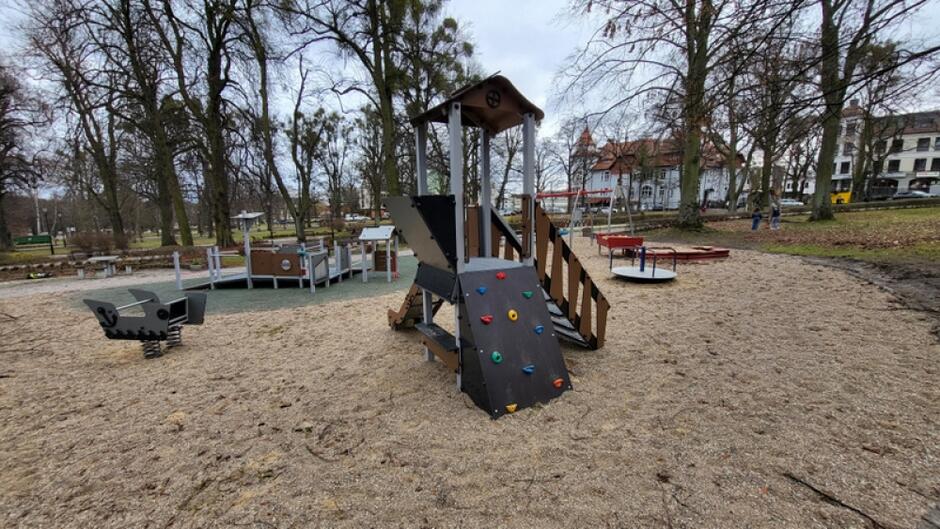 na zdjęciu plac zabaw, widać piaskową nawierzchnię i drewnianą ciemną konstrukcję do zabawy dla dzieci, po której można chodzić i ślizgać się