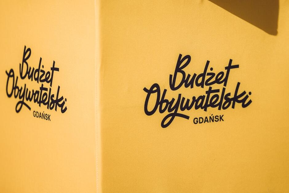 Papierowa ulotka żółtego koloru, nieco zgięta w pionie. Po lewej i po prawej stronie od zgięcia widzimy dwa drukowane napisy Budżet Obywatelski Gdańsk