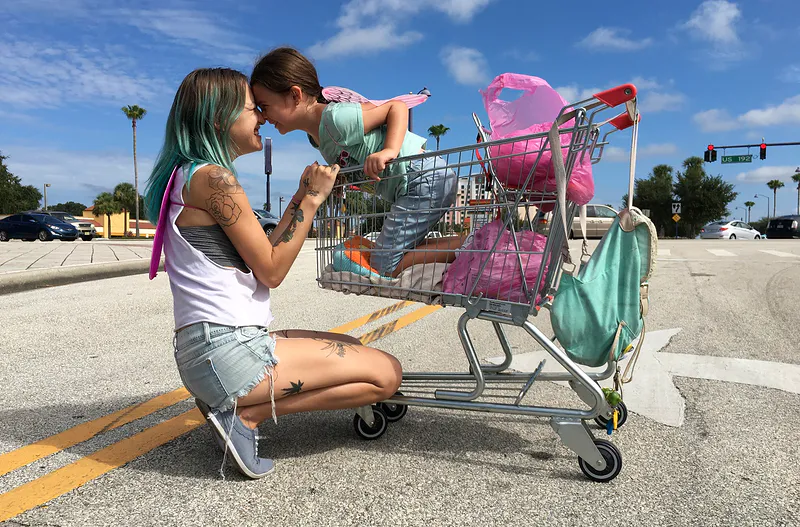 dziewczynka w wózku sklepowym, przy nim klęczy młoda dziewczyna