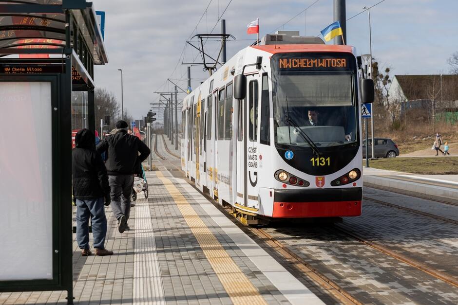 Zdjęcie białego tramwaju, który podjeżdża do przystanku, na którym stoi kilka osób. Tramwaj jest po prawej, przystanek - po lewej