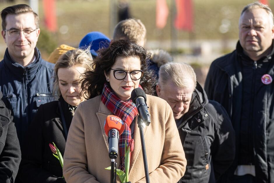 Kobieta w okularach w ciemnych włosach stoi przed mikrofonem. Obok kilka innych osób