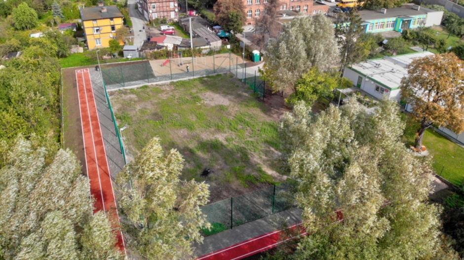 zdjęcie z drona, widać ogrodzony gruntowy tereny pokryty trawą, po lewej jest bieżnia lekkoatletyczna, po prawej widać niewysokie zabudowania