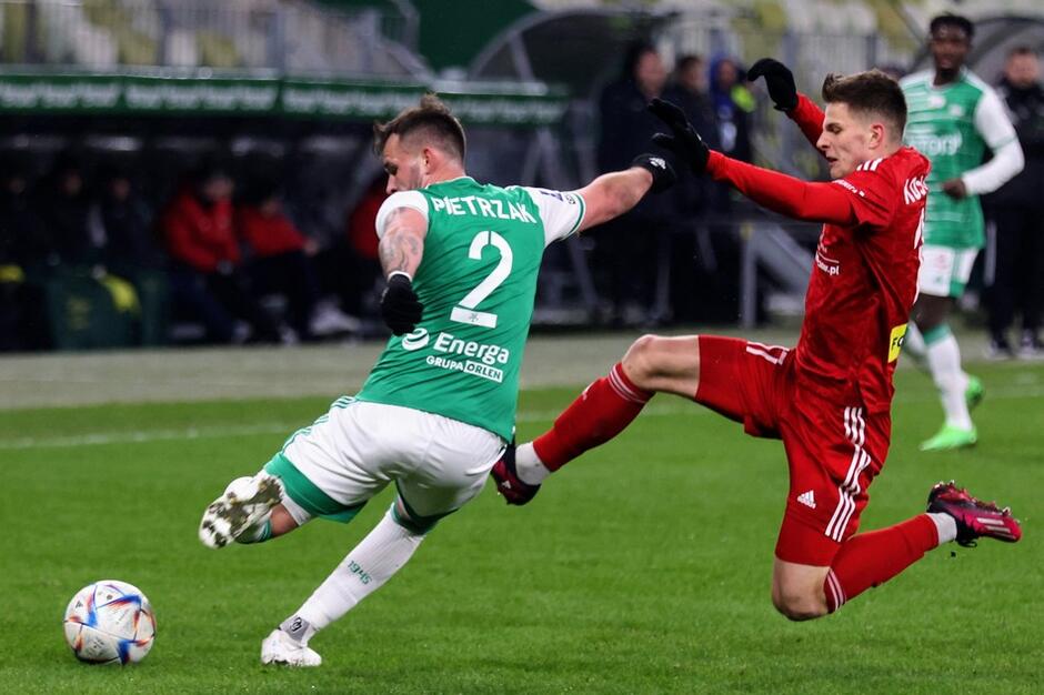 Piłkarz w zielonej koszulce przy piłce, blokuje zawodnik w czerwonym stroju