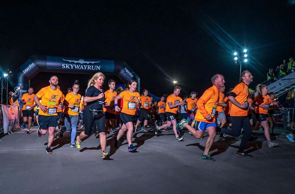 biegacze nocą w pomarańczowych koszulkach tuż po starcie, biegną noc po płycie lotniska