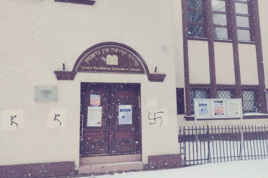 Widok ogólny wejścia do synagogi we Wrzeszczu. Po prawej od brązowych drzwi jest swastyka, po lewej - widzimy dwie litery K, wykonane na tej samej wysokości muru. Wszystkie znaki zrobiono czarnym sprayem