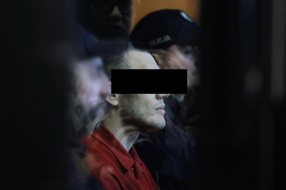 Twarz młodego człowieka w czerwonym więziennym drelichu - pokazana z blika. Ma oczy zasłonięte czarnym prostokątem 