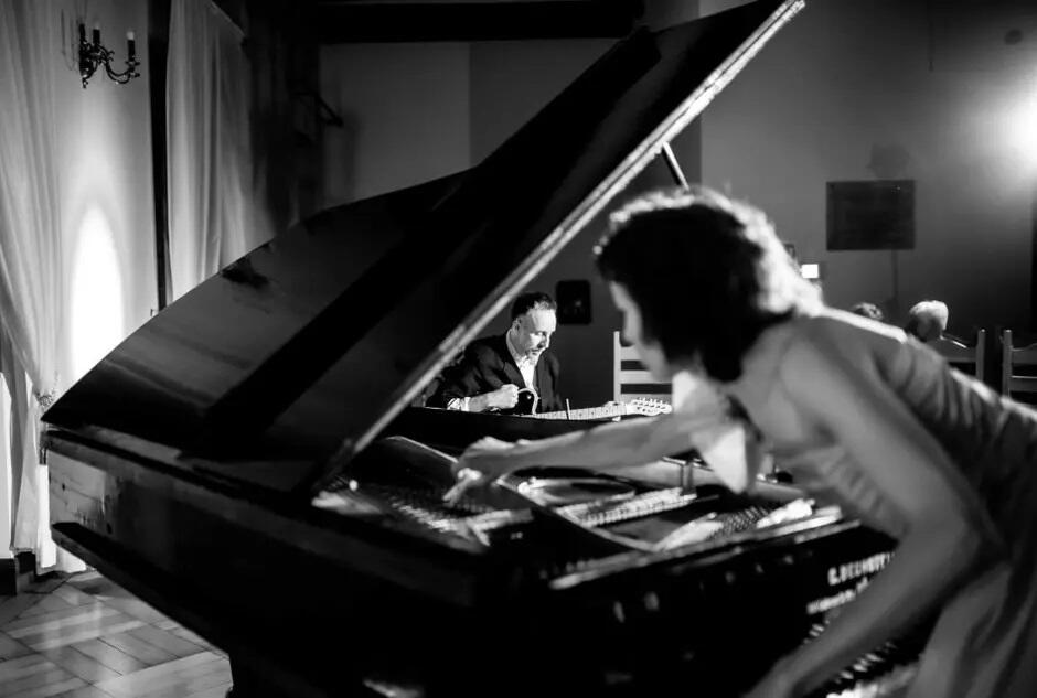 Na czarno białym zdjęciu widać fortepian i młodą kobietę, która pochyla się nad klawiaturą. W tle grającego na gitarze mężczyznę.
